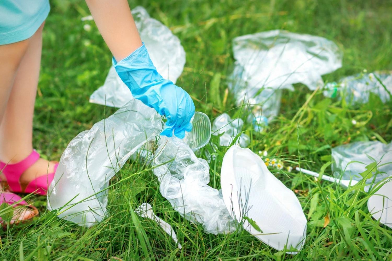 Child wearing a blue glove picking up plastic waste from grass during a park cleanup